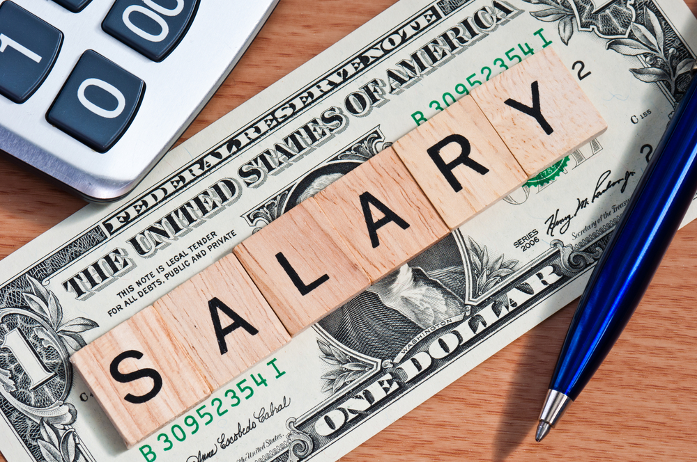 A US dollar bill with scrabble letters spelling out salary sitting atop a table